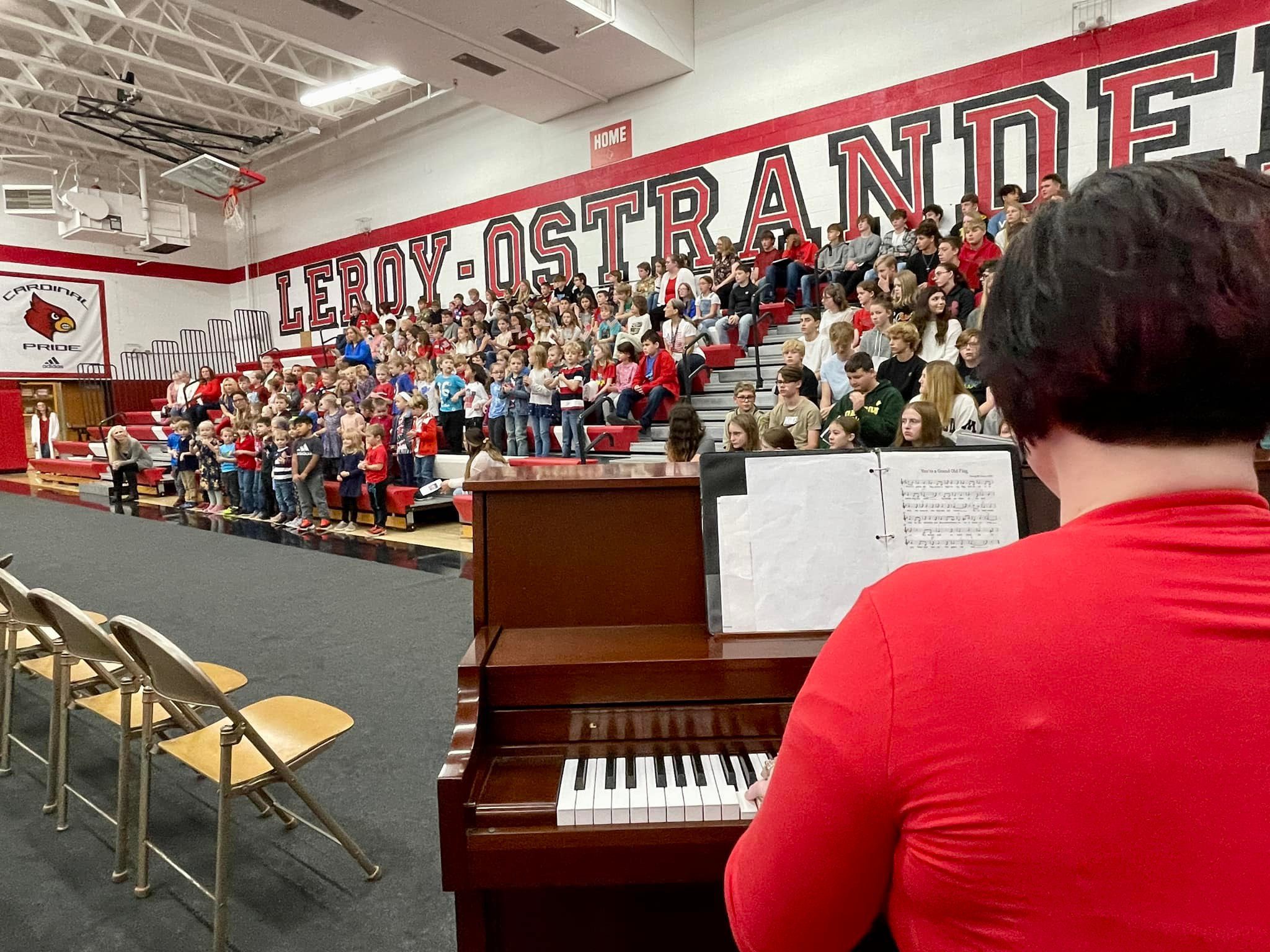 A woman playing the piano with students singing in the background.