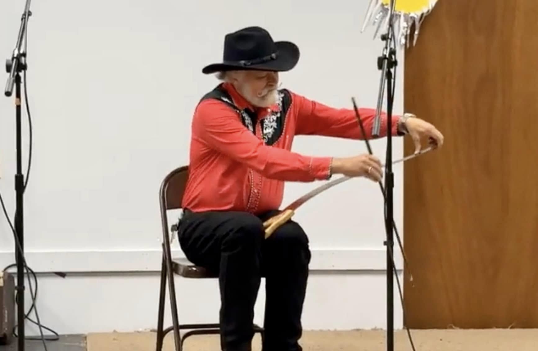 A man sitting on a chair making music with his saw.